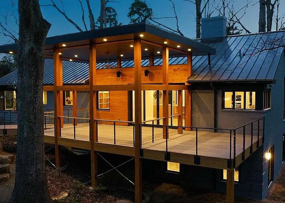 Modern wooden house exterior with deck, dim lighting, and surrounding trees at dusk.