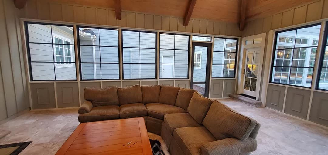 Cozy sunroom with large windows, sectional sofa, and wooden coffee table. Bright and inviting space.