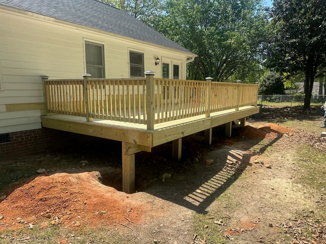Newly constructed wooden deck with railings, elevated above ground beside a house.