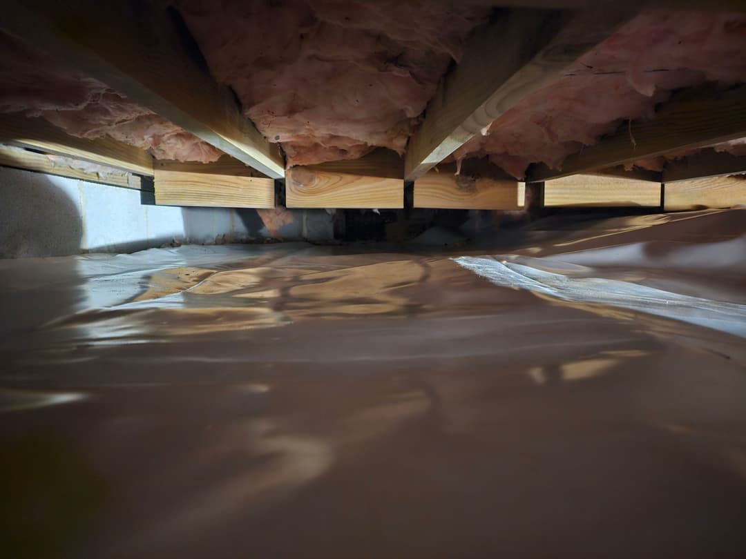 View of a crawl space with wooden beams and plastic sheeting, highlighting insulation and moisture control.