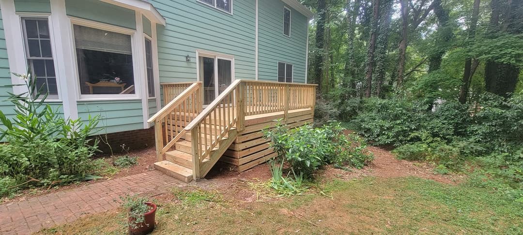 New wooden deck with ramp beside a green house, surrounded by lush trees and landscaping.