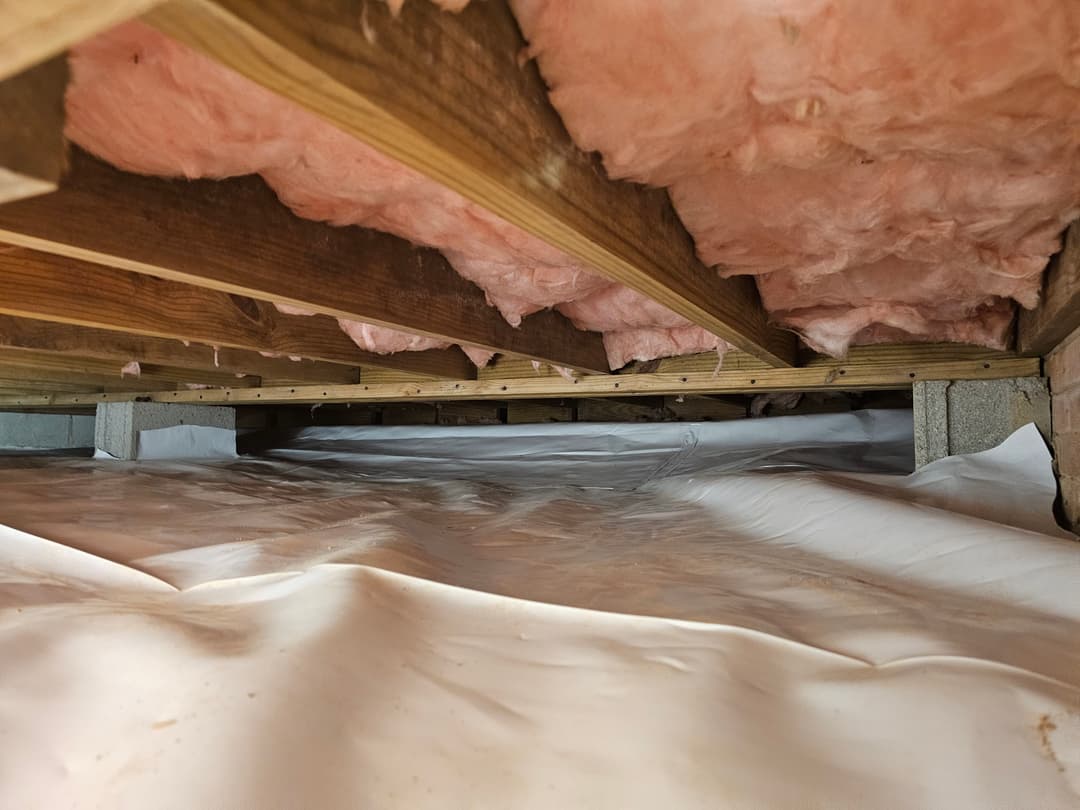 Crawl space with pink insulation and vapor barrier on the floor beneath wooden beams.