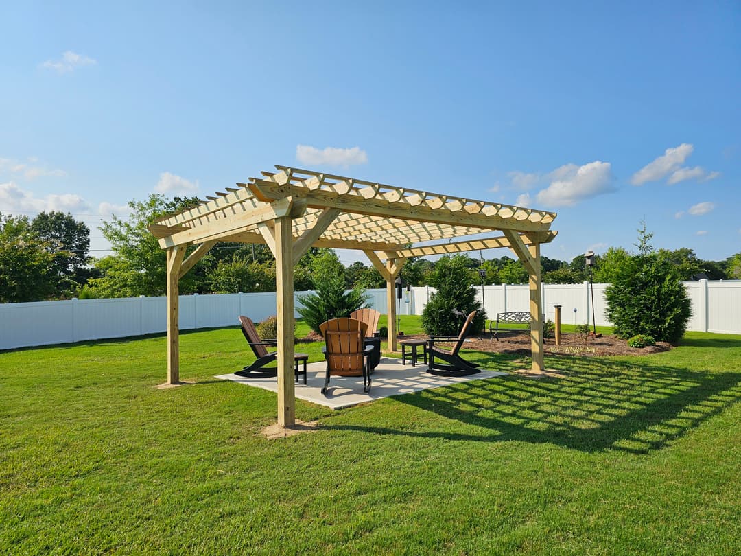 Wooden pergola with chairs in a landscaped backyard under a blue sky.