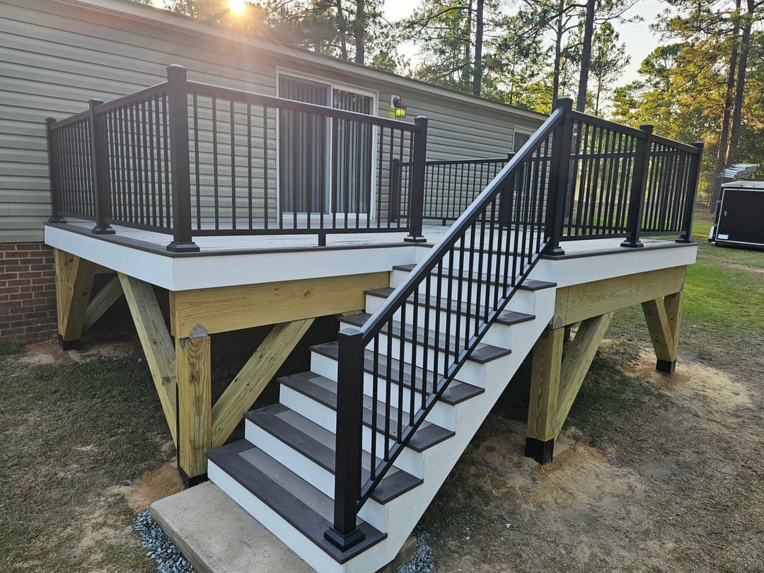 Modern elevated deck with black railing and stairs, surrounded by trees and sunlight.