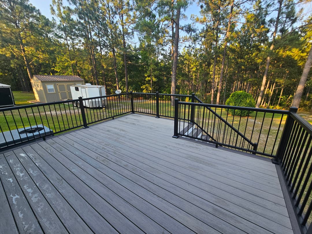 Spacious wooden deck with black railings surrounded by pine trees and outdoor storage shed.