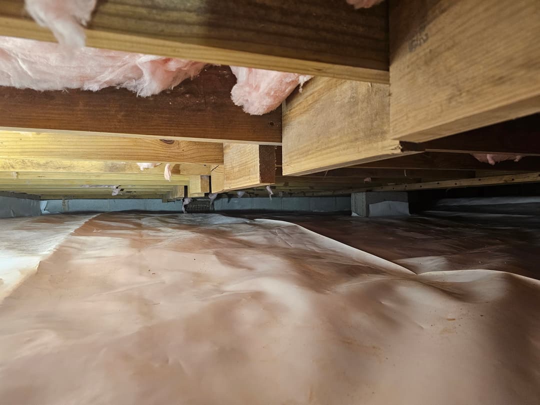 View of a crawl space with wooden beams and plastic sheeting on the ground.